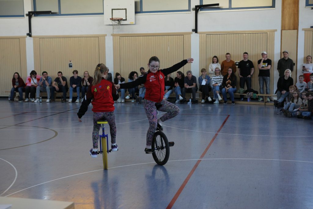 Foto zeigt in der Pause eine Vorführung von zwei Einrad Mädchen auf dem Einrad in der Halle, an der Hand haltend und im Kreis fahrend.