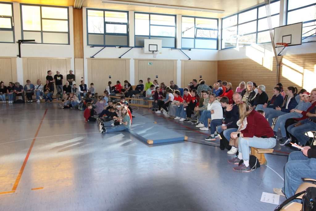 Foto zeigt die Turnhalle und die Teilnehmer sitzend auf Stühlen und Bänken mitten in der Halle verteilt.