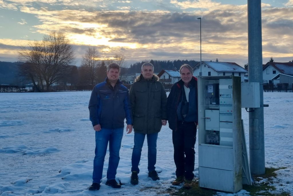 Foto zeigt vor einem Schaltschrank Elektronik der geöffnet ist und auf dem hinteren Trainingsplatz im Sonnenuntergang den Vertriebsmitarbeiter der Firma LED Sport, dem Vize-Präsidenten TSV und dem TSV Präsidenten.
