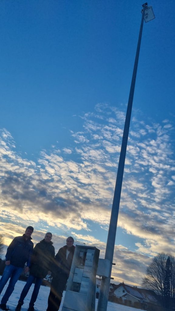 Foto zeigt vor einem Schaltschrank Elektronik der geöffnet ist und auf dem hinteren Trainingsplatz im Sonnenuntergang den Vertriebsmitarbeiter der Firma LED Sport, dem Vize-Präsidenten TSV und dem TSV Präsidenten. Hier ist noch der ganze Mast in voller Höhe zu sehen.